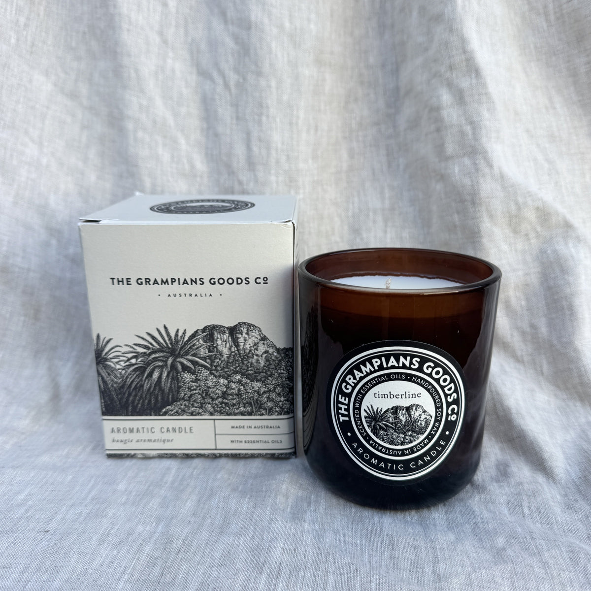 An amber glass jar candle next to its white cardboard packaging box, both featuring the brand name 'The Grampians Goods Co' and a black-and-white illustration of mountains and plants, set against a textured light gray fabric background.