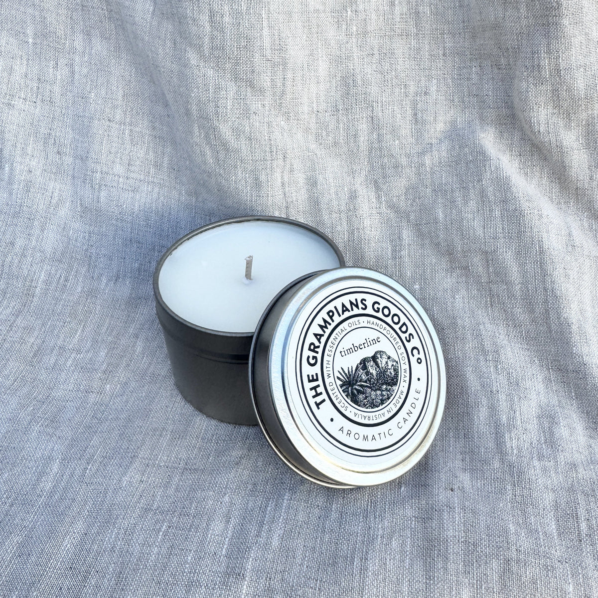 A black metal candle tin with a white wick, showing white wax inside, next to its silver lid which features a circular logo for 'The Grimpians Goose Co.' on a textured gray linen background.
