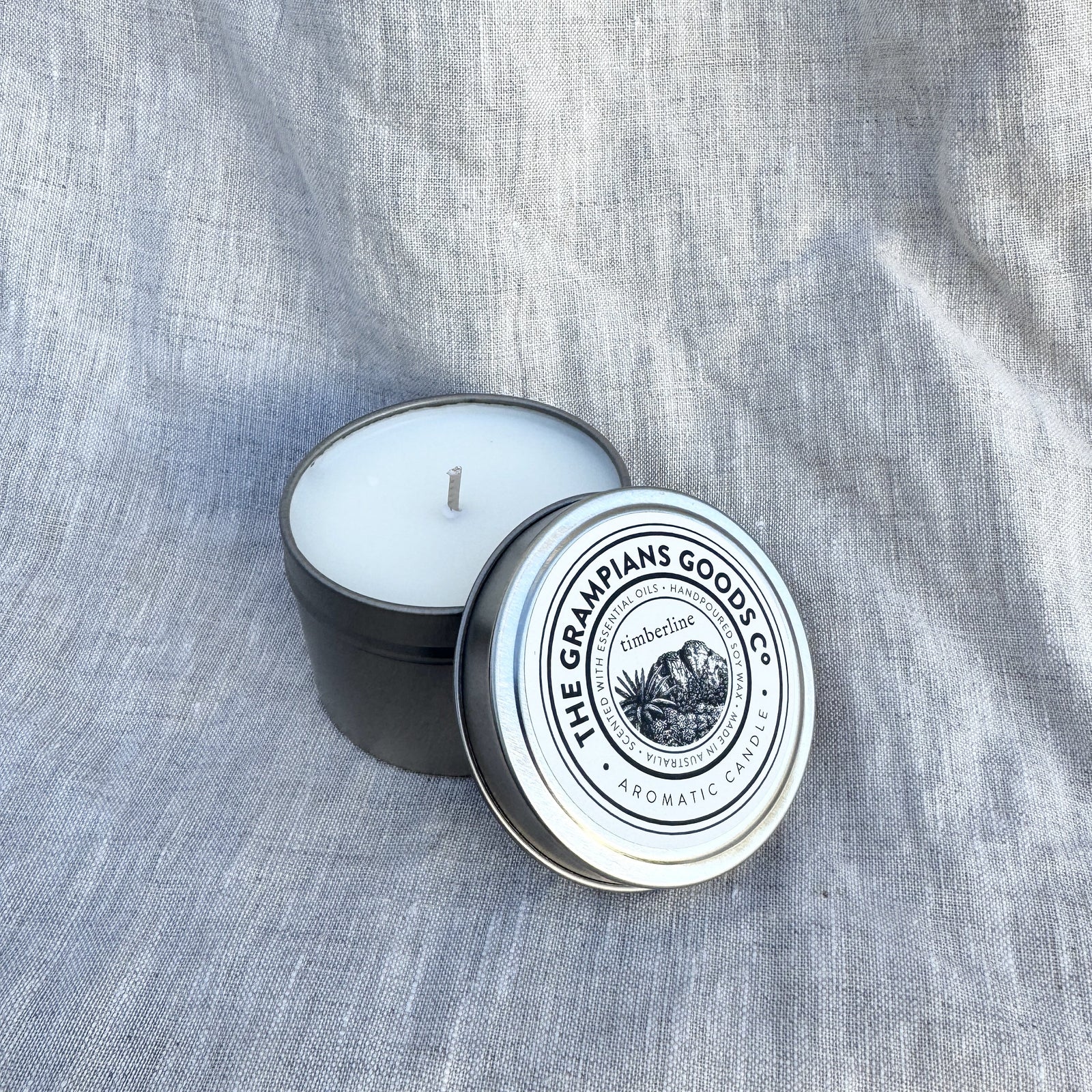 A black metal candle tin with a white wick, showing white wax inside, next to its silver lid which features a circular logo for 'The Grimpians Goose Co.' on a textured gray linen background.